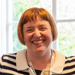 A smiling person with short brown hair wearing a striped shirt and a lanyard stands indoors in front of a sunlit window.