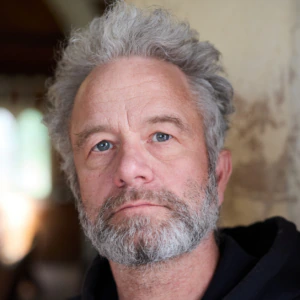 A middle-aged man with gray hair and a beard, wearing a black hoodie, stands indoors against a light-colored wall, looking directly at the camera with a neutral expression.