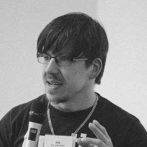 A person holding a microphone speaks while gesturing with their other hand. They wear glasses, a T-shirt, and a name tag.