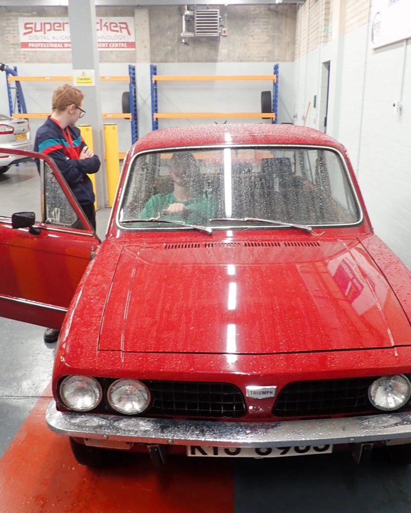 Two people work on a red car, one sitting inside and one standing outside with the door open