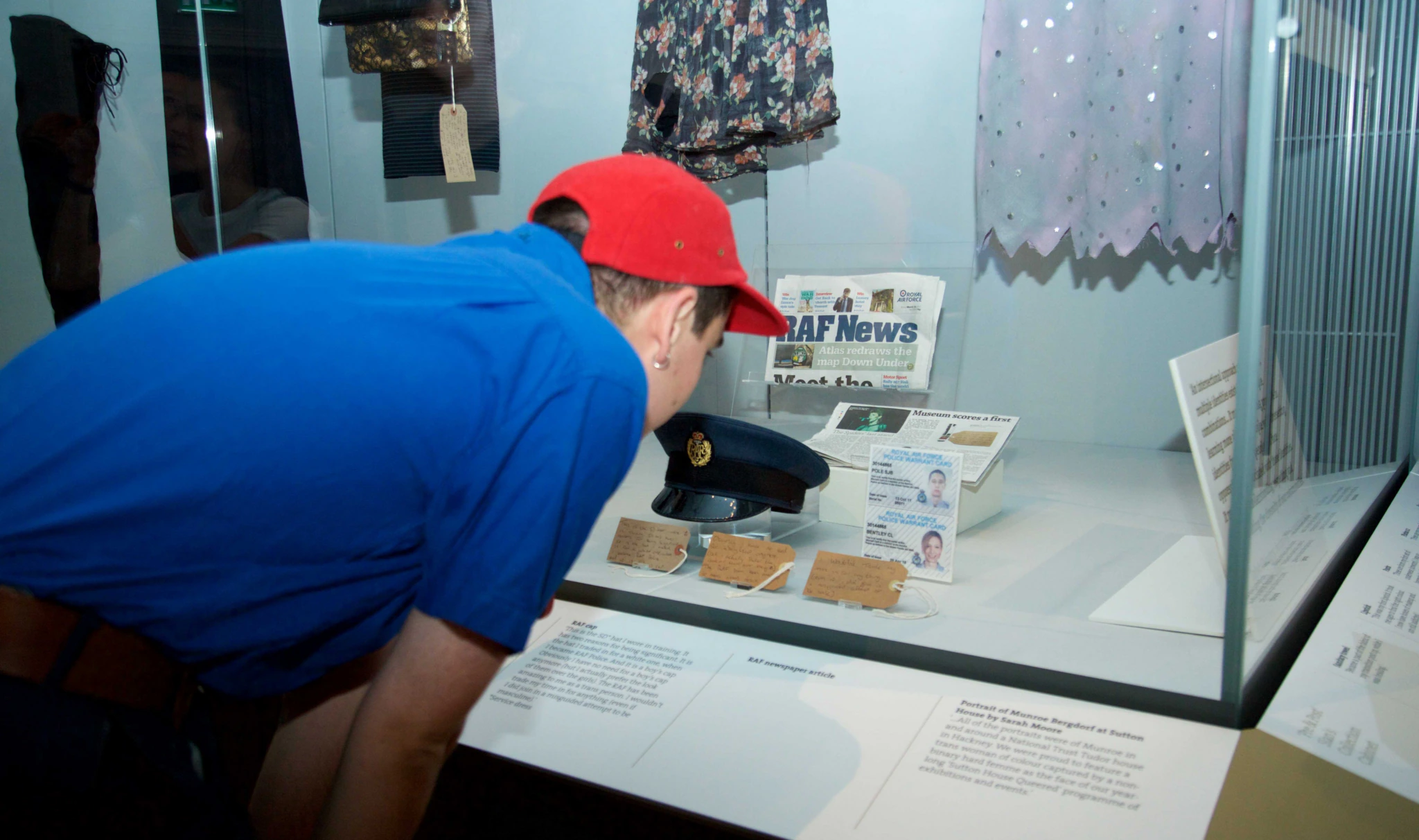 A person in a blue t shirt and red cap bends over to look into a display case. Inside the display case, is a militery-style hat, photo ID cards and several brown object labels.