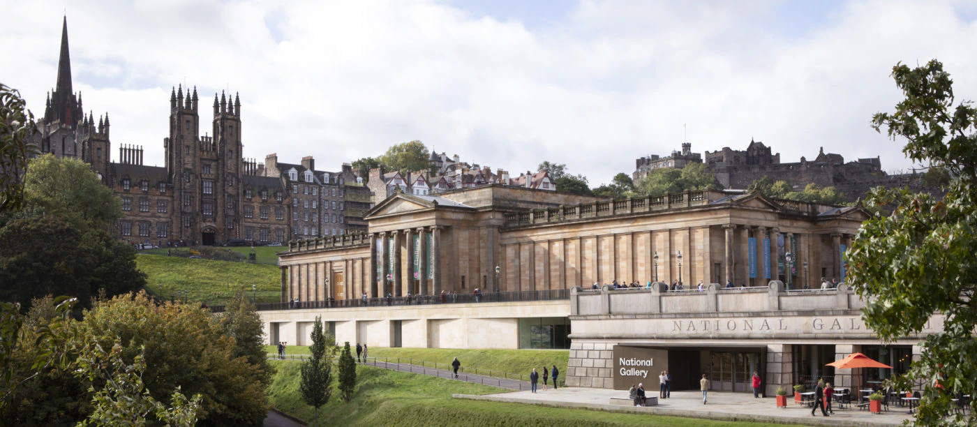 A huge, and grand, columned museum overlooking stepped lawn with a meandering path through it set against the backdrop of Edinburgh's spiky skyline and a cloudy daylit sky