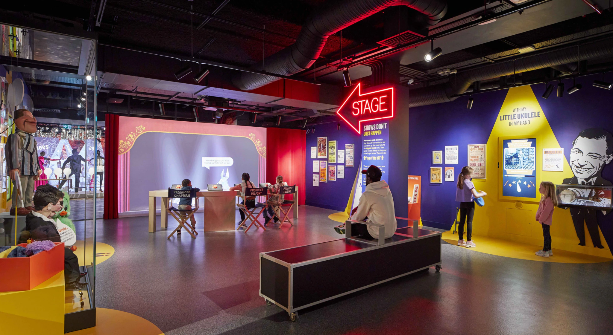 Darkened gallery with lit sign saying "Stage" in an arrow shape and people on directors chairs in front of a screen