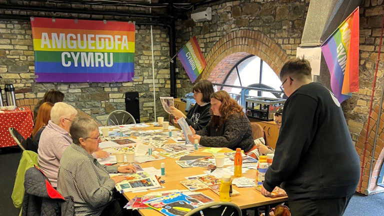 People sit around a table and take part in an engagement activity - pride flags in the background read Amgueddfa Cymru