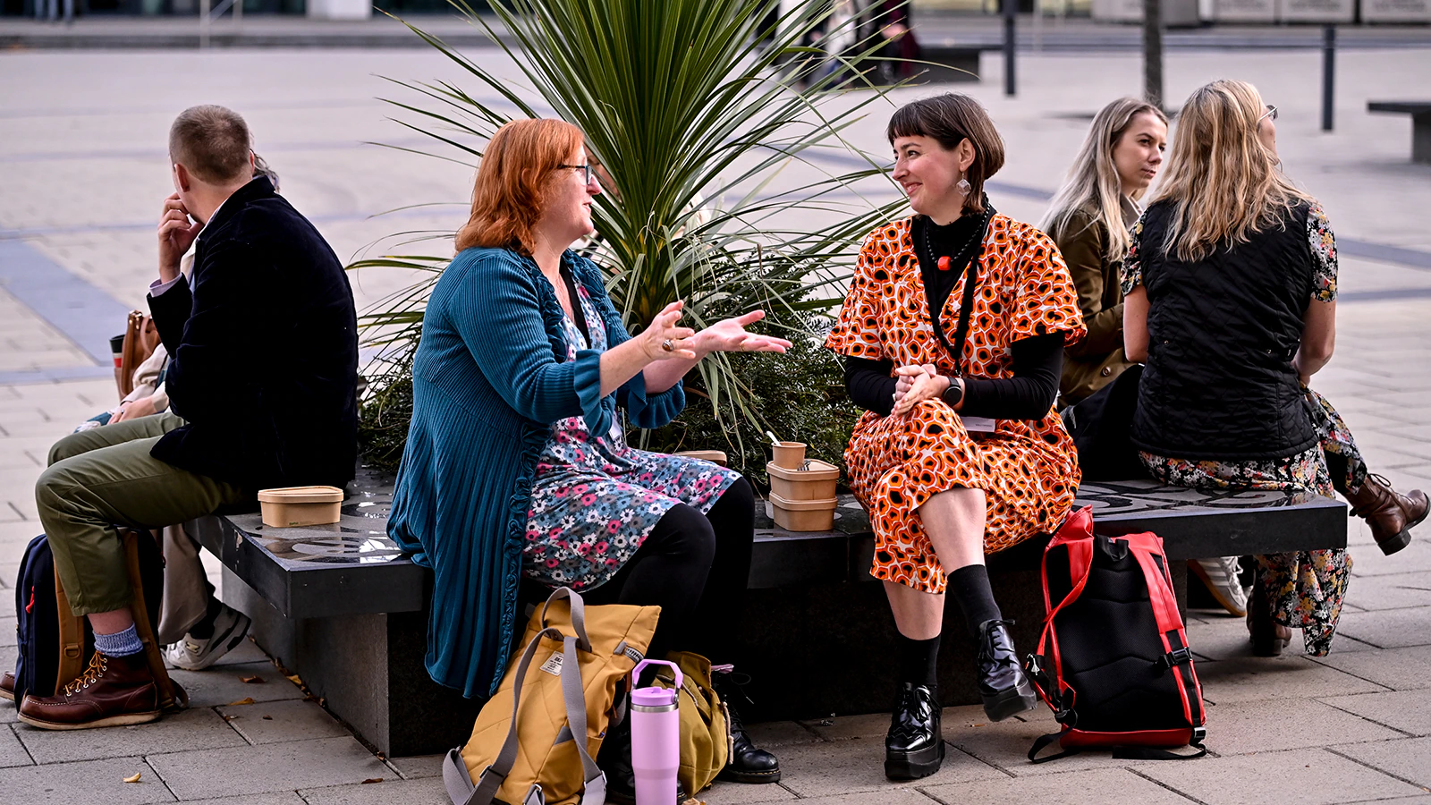 An array of people sit on a circular bench outdoors and gesture as they speak to one another