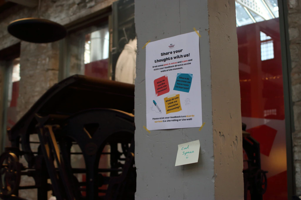 A poster on a grey pillar invites people to Share your thoughts with us! It features coloured speech bubbles and a QR code. A sticky note below reads: Cool space. Background shows an industrial setting with a large machine.