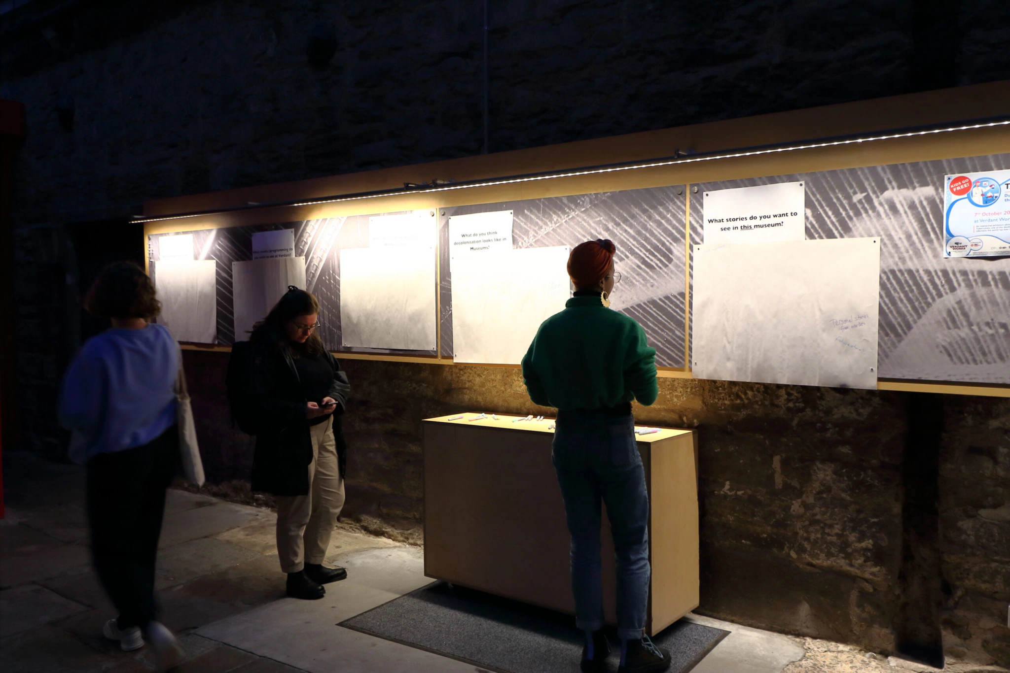 Three people stand at a dimly lit exhibit with boards featuring small posters and notes. One person is writing, another is looking at their phone, and the third person is observing. A wooden stand is in the centre.