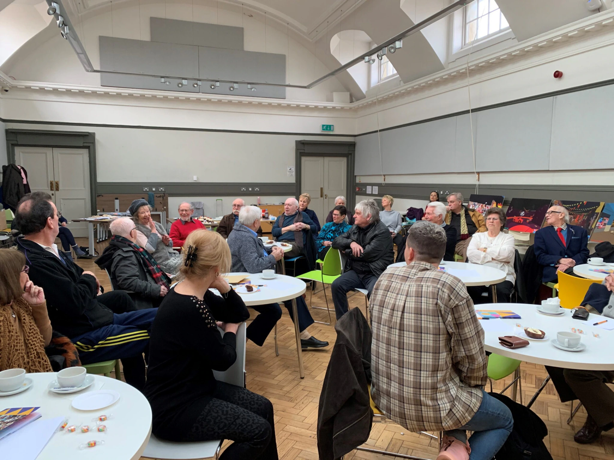 A meeting of the Showtown Performers' Network in 2020. A group of people sit in a room with a high ceiling, gathered around tables for a discussion or meeting. The space is well-lit, with windows and artwork on the walls. Cups and papers are scattered on the tables.