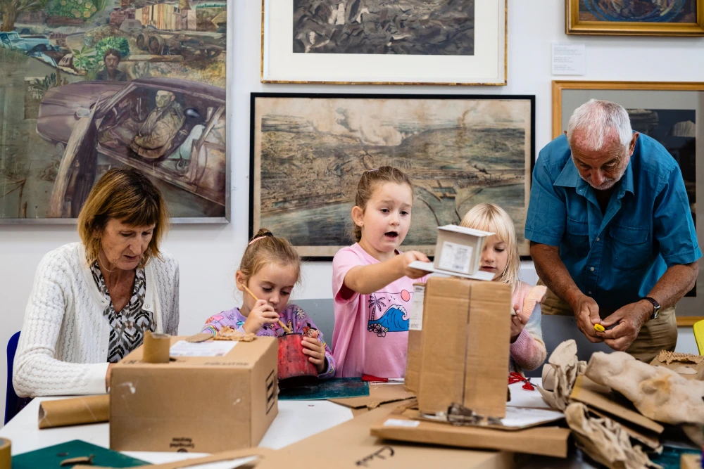 A workshop at Glynn Vivian Art Gallery, Swansea. Adults and children engaged in an arts and crafts activity, assembling materials such as cardboard and paper. The table is strewn with various art supplies, and paintings hang on the wall in the background.