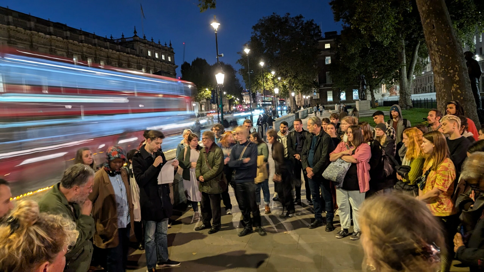 A Museum of Homelessness vigil for those who died while homeless in 2023. A group of people gathered outdoors at night near a tree, listening to a speaker holding a paper. A double-decker bus, captured in motion blur, passes by. Streetlights and a historic building are visible in the background.