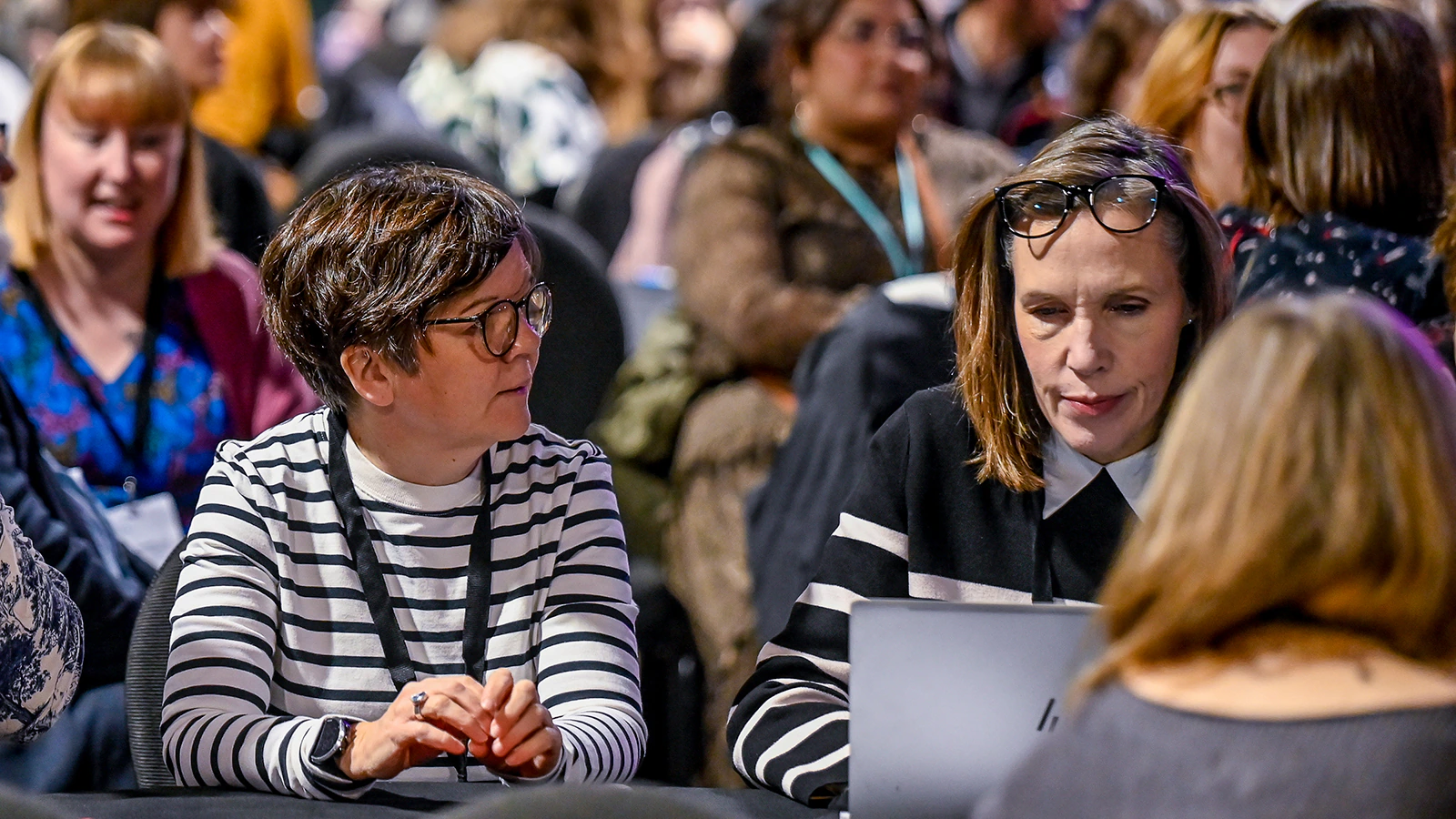 Two women are sitting at a table in a crowded room, engaged in a discussion. One is wearing a striped shirt and glasses, the other a black and white outfit