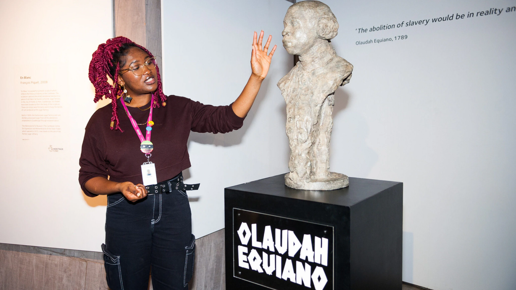 National Museums Liverpool. A person with red braids and glasses gestures towards a sculpture on display. The sculpture is of Olaudah Equiano, with his name labelled on the pedestal.