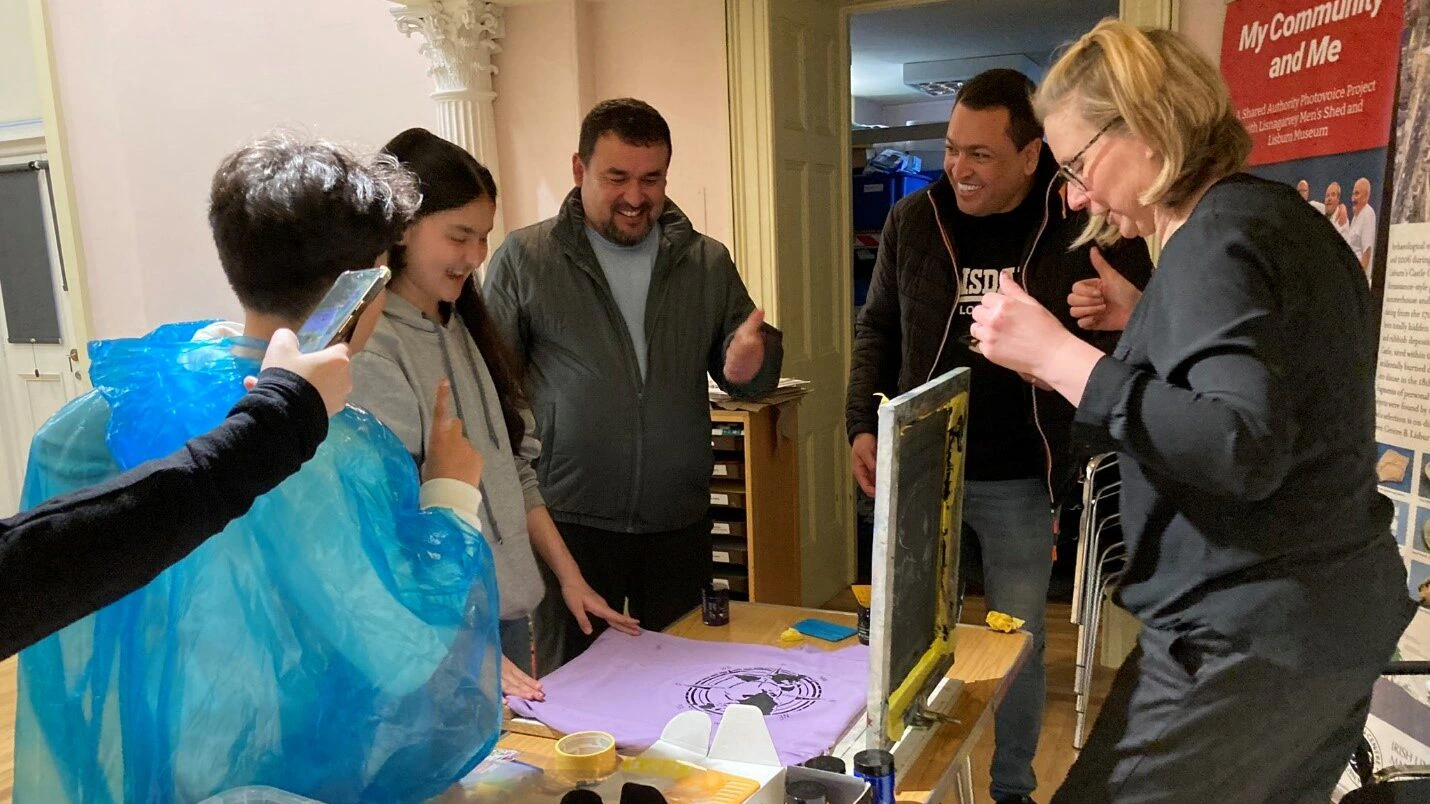 Lisburn Museum, depicting an artist session with the design of screen prints chosen by Syrian and Afghan participants. A group of people are gathered around a table with screen printing equipment. A woman is demonstrating printing on fabric while others watch and smile. Art supplies and a colourful sign, My Community and Me, are visible in the background.