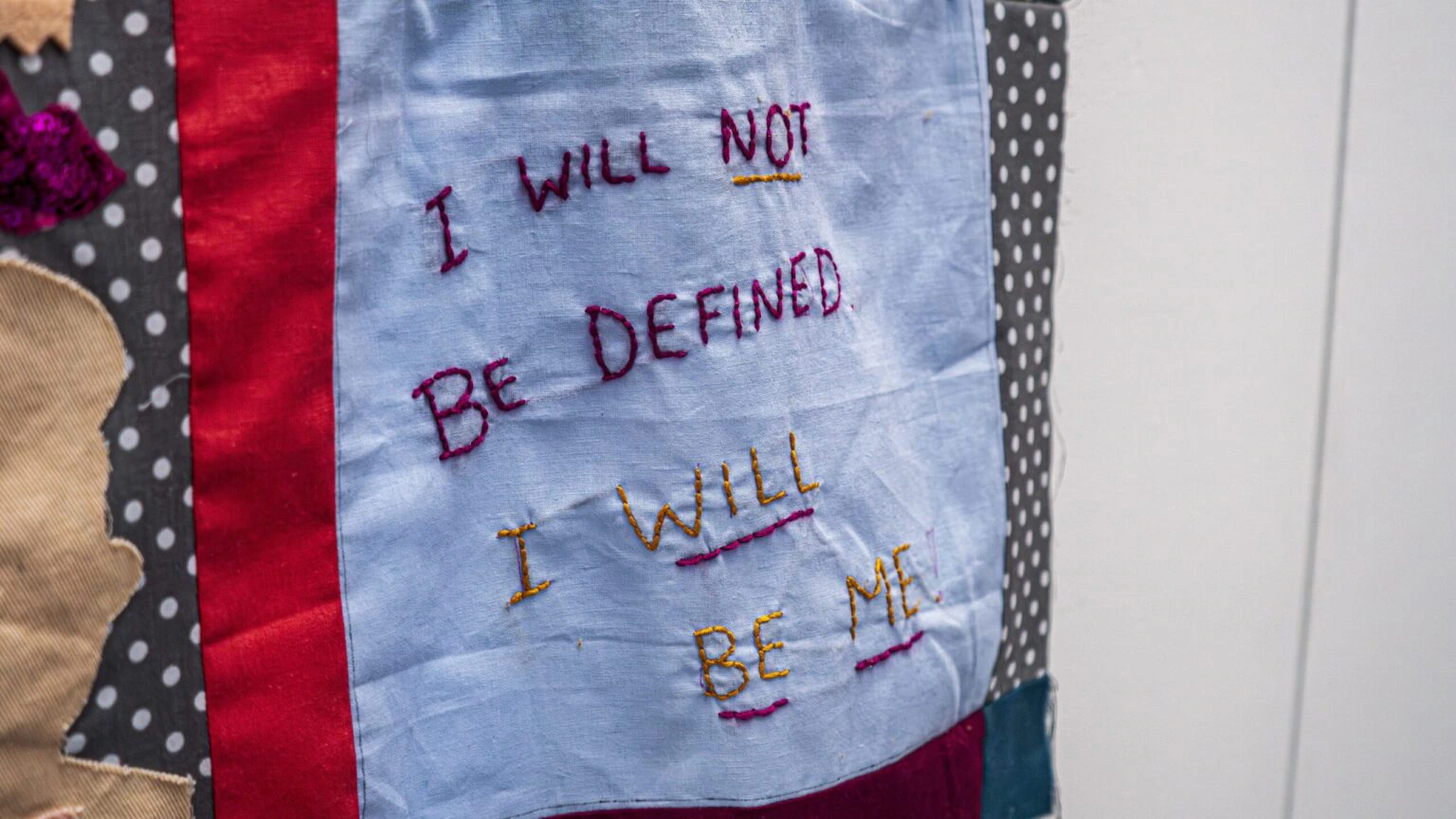 Close-up of a fabric patch with colorful embroidery. Text reads, I will not be defined, I will be me, on a blue background, bordered by polka dot and red fabric.