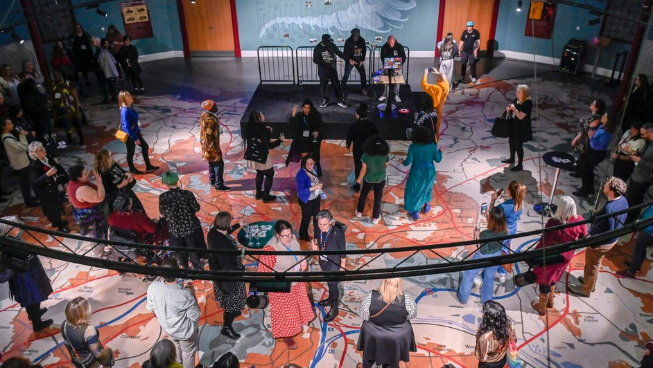 A group of people gather at a lively indoor event. They stand and mingle on a floor decorated with map-like patterns under a large installation. Some are interacting or watching a couple of performers on a small stage. The atmosphere is festive and social.