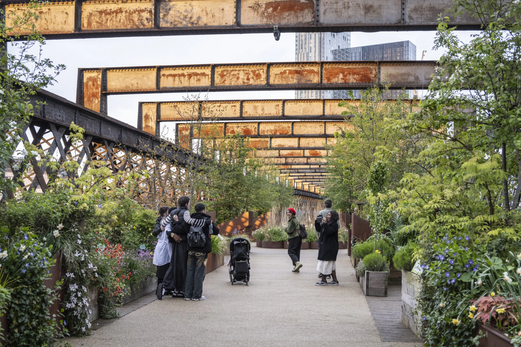 People walk and take photos on a plant-lined urban walkway with rusty iron beams overhead. Some people are gathered near a stroller, surrounded by lush greenery and city buildings in the background.