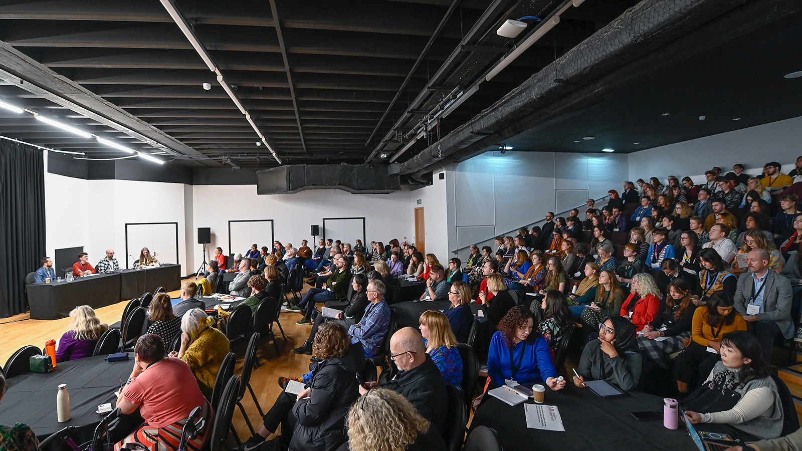 A large group of people sit in a modern auditorium, attentively listening to a panel of speakers at the front
