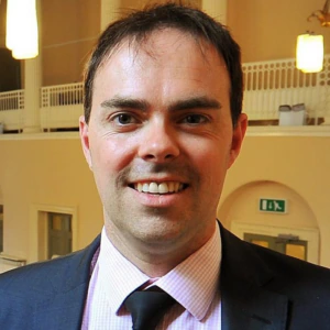 A man in a suit and tie smiles at the camera in a well-lit room with cream walls, hanging lights, and a balcony with white railing in the background.
