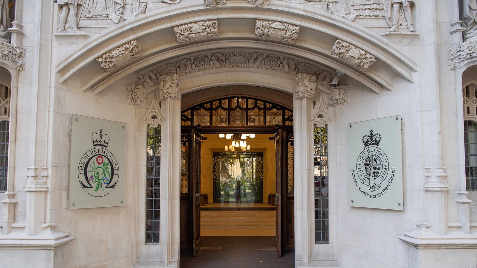 Ornate stone entrance to a historic building with two large plaques, each displaying a crest and text. Glass doors and wood detailing are visible inside, with a modern reception area and hanging lights.