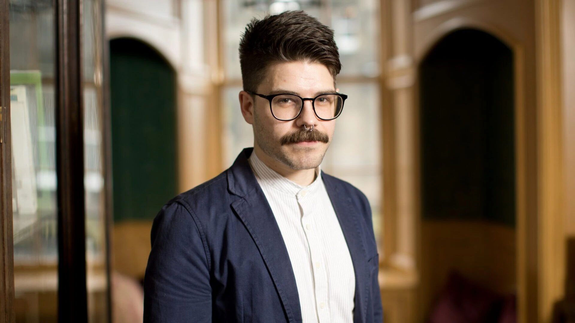 A person with a mustache, short brown hair, and glasses is wearing a blue blazer over a white shirt, standing indoors in a wood-paneled room with arched alcoves and soft lighting.