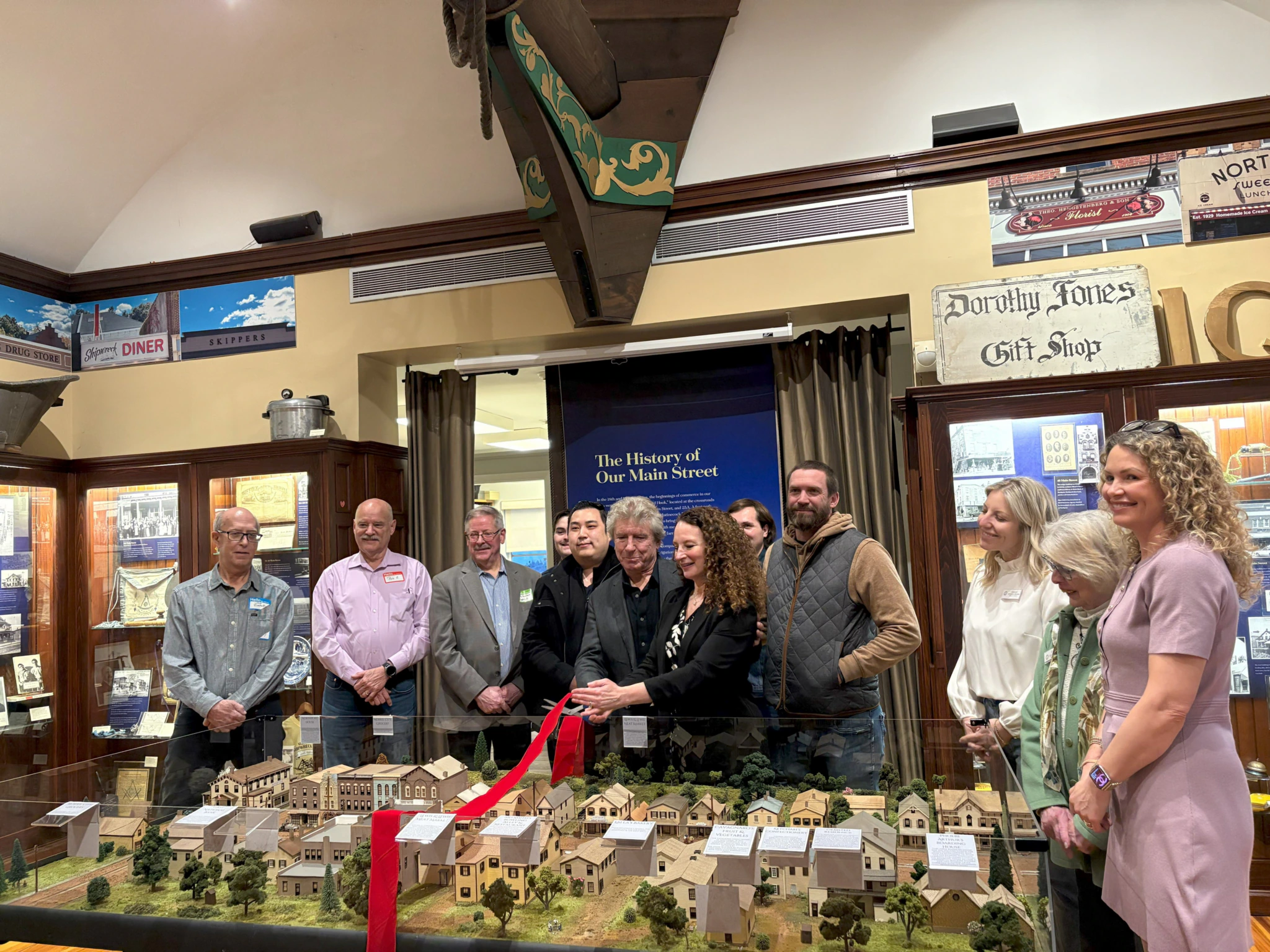 A group of people stands behind a display model of a town, some holding large scissors about to cut a red ribbon. They are in a museum with historic signs and photos on the walls behind them.