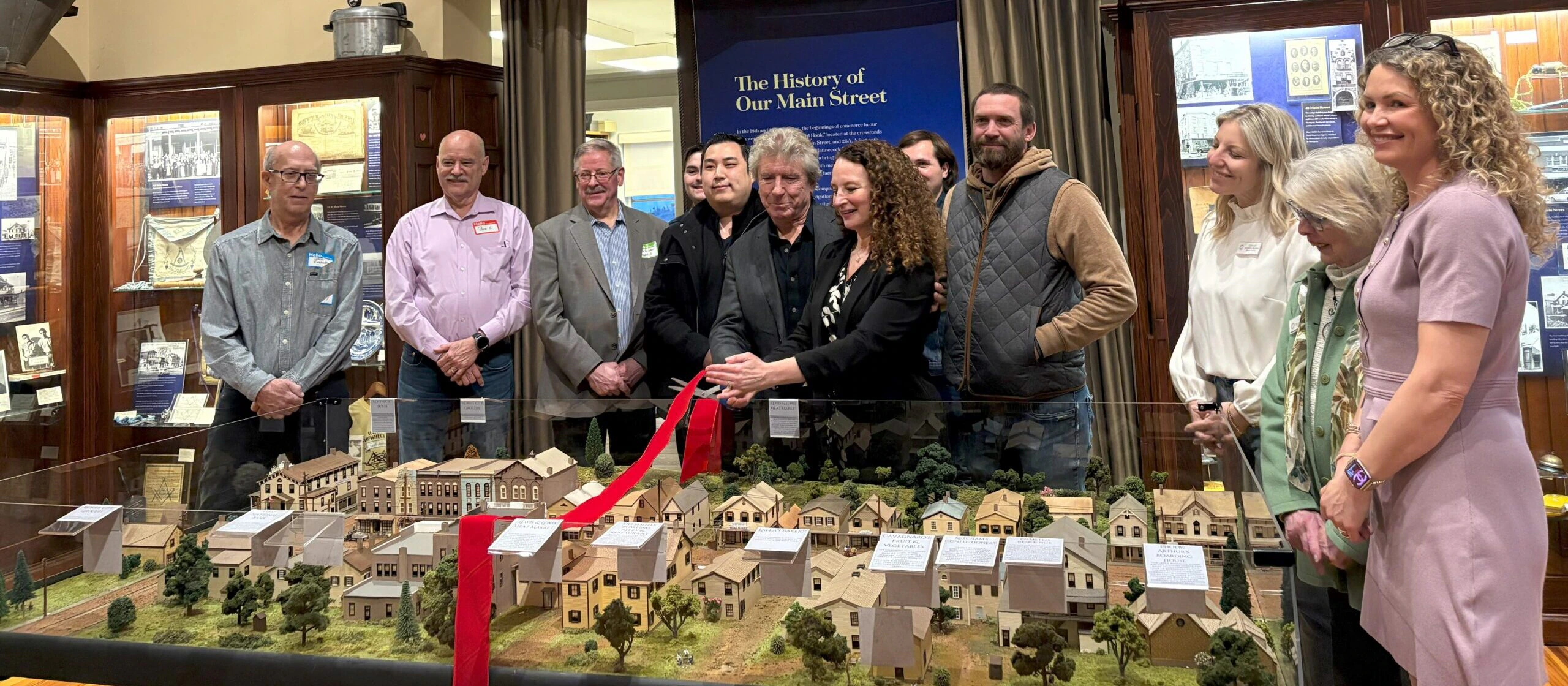 A group of people stands behind a display model of a town, some holding large scissors about to cut a red ribbon. They are in a museum with historic signs and photos on the walls behind them.