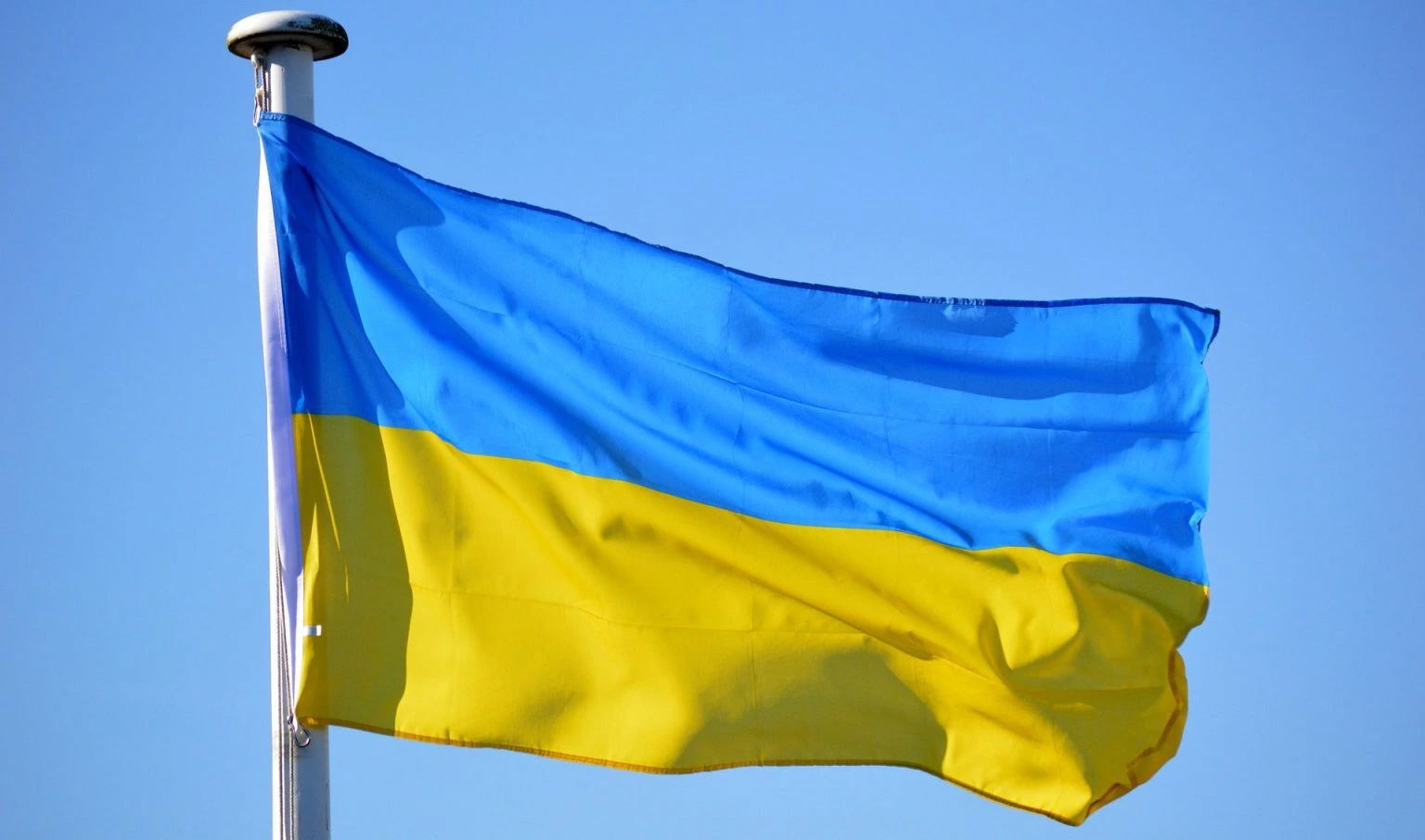 The Ukrainian flag, with blue on the top half and yellow on the bottom half, waves on a flagpole against a clear blue sky.