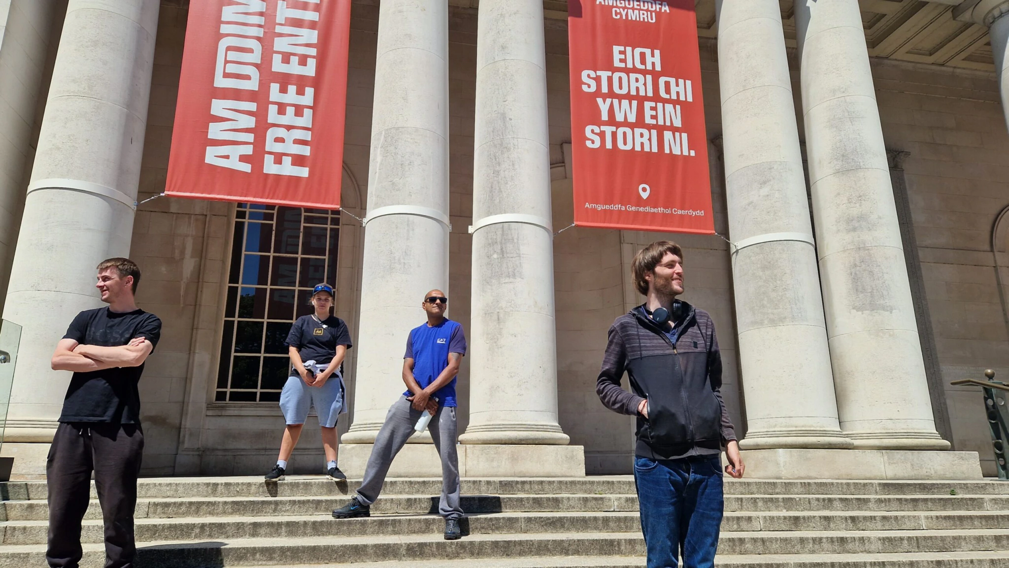 Four people stand on the steps of a building with tall columns. Two large red banners hang above them, one reading FREE ENTRY and the other with Welsh text. The weather is sunny and bright.