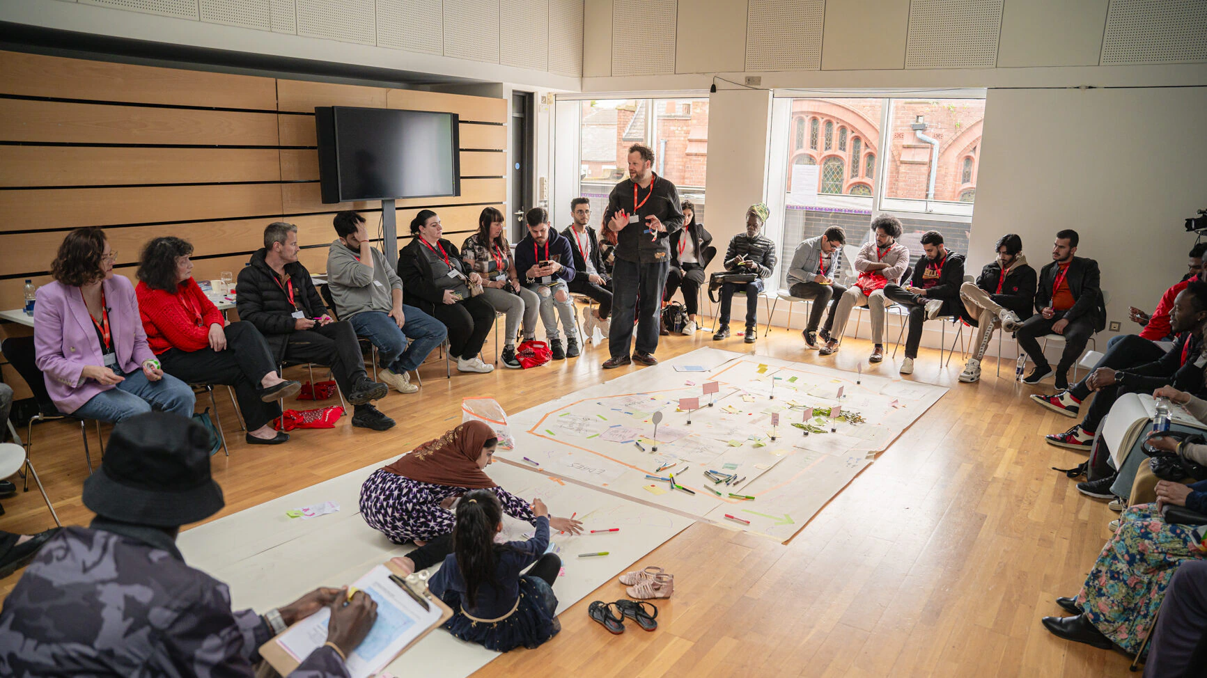 A group of people sit in a large circle in a bright room, watching a standing man speak