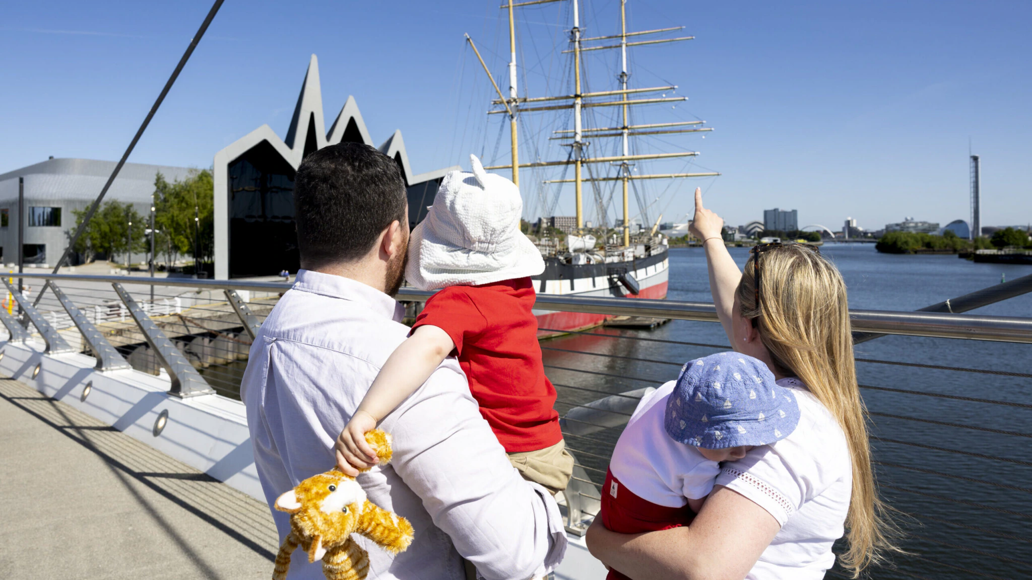 A family with two young children stands on a bridge, looking at a large sailing ship docked on the river. One child holds a stuffed toy, while an adult points toward the ship under a clear blue sky.
