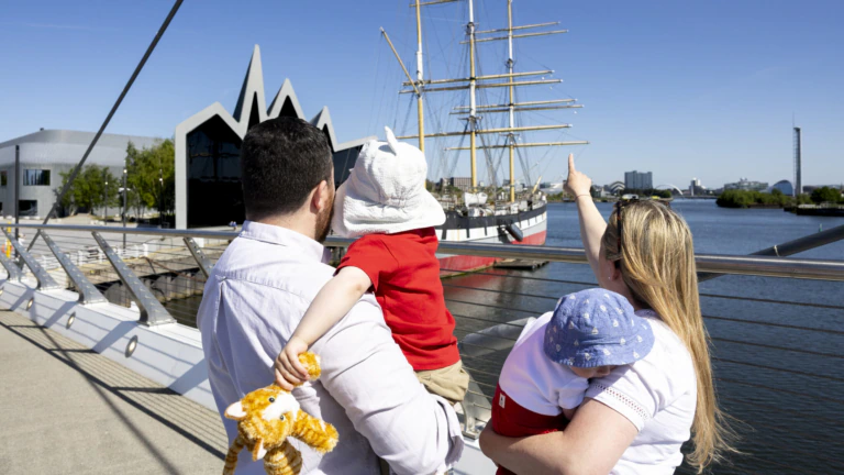 A family with two young children stands on a bridge, looking at a large sailing ship docked on the river. One child holds a stuffed toy, while an adult points toward the ship under a clear blue sky.