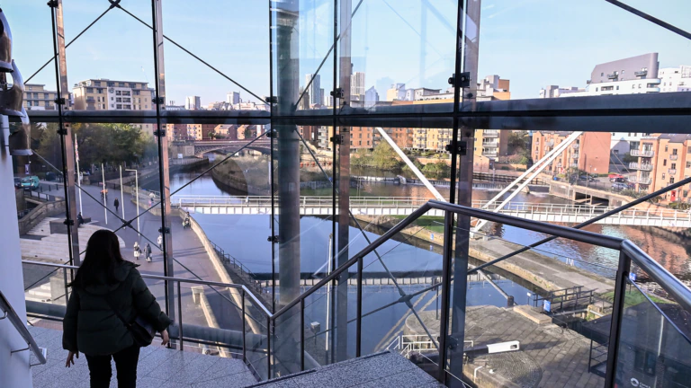 A person walks down indoor stairs by large glass windows overlooking a river, bridges, and modern city buildings under a clear sky.