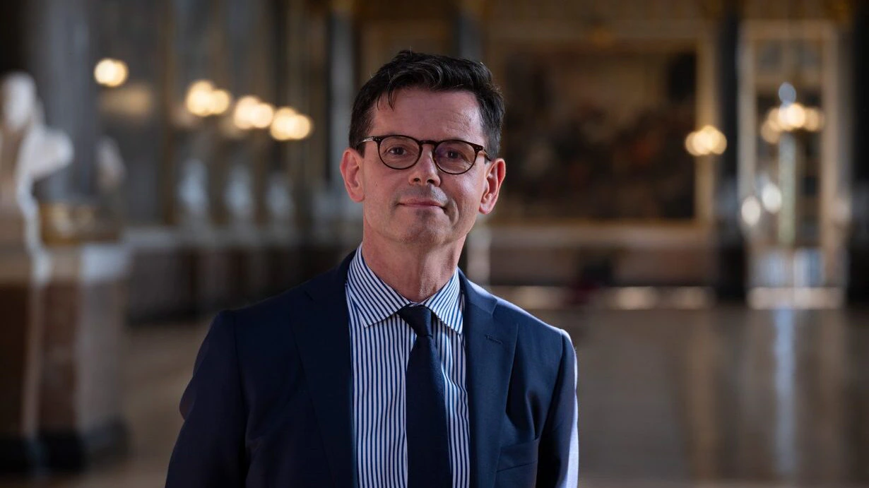 A man in a suit and glasses stands in a large, elegant hall with high ceilings, soft lighting, and blurred statues and paintings in the background.