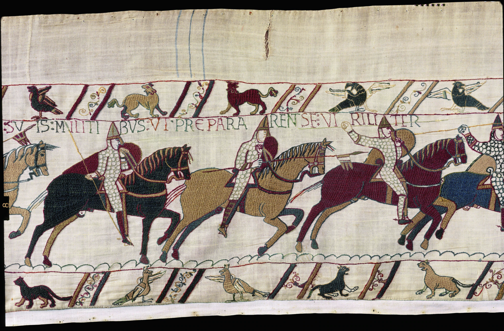 A section of the Bayeux Tapestry shows armored soldiers on horseback, carrying spears and shields, with animals and birds above and Latin text embroidered at the top.