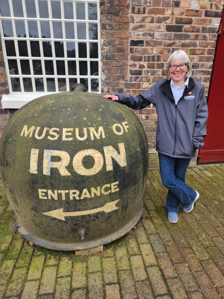 A smiling woman in glasses and a gray jacket stands next to a large round stone sign reading Museum of Iron Entrance with arrows, outside a brick building with a window and a red door.