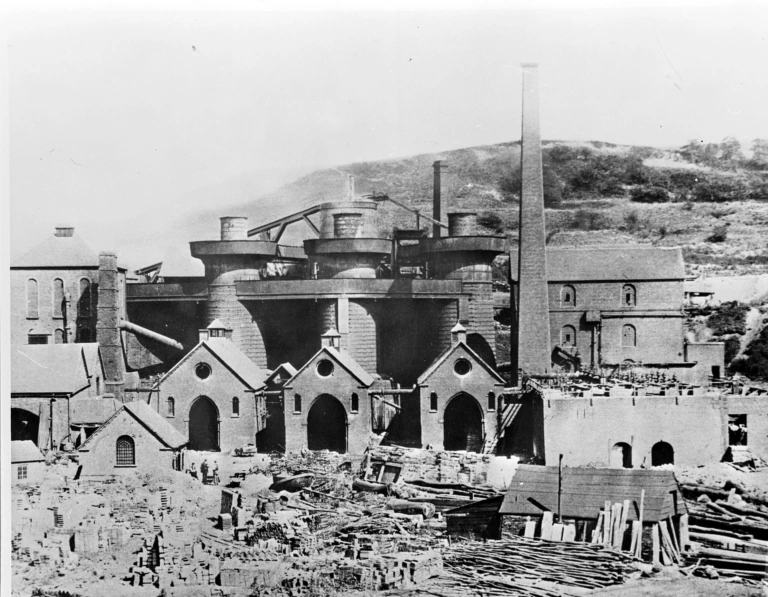 Historic black-and-white photo of an industrial ironworks with tall chimneys, large furnaces, and several brick buildings; scattered materials and machinery are visible in the yard in front. Hills rise in the background.