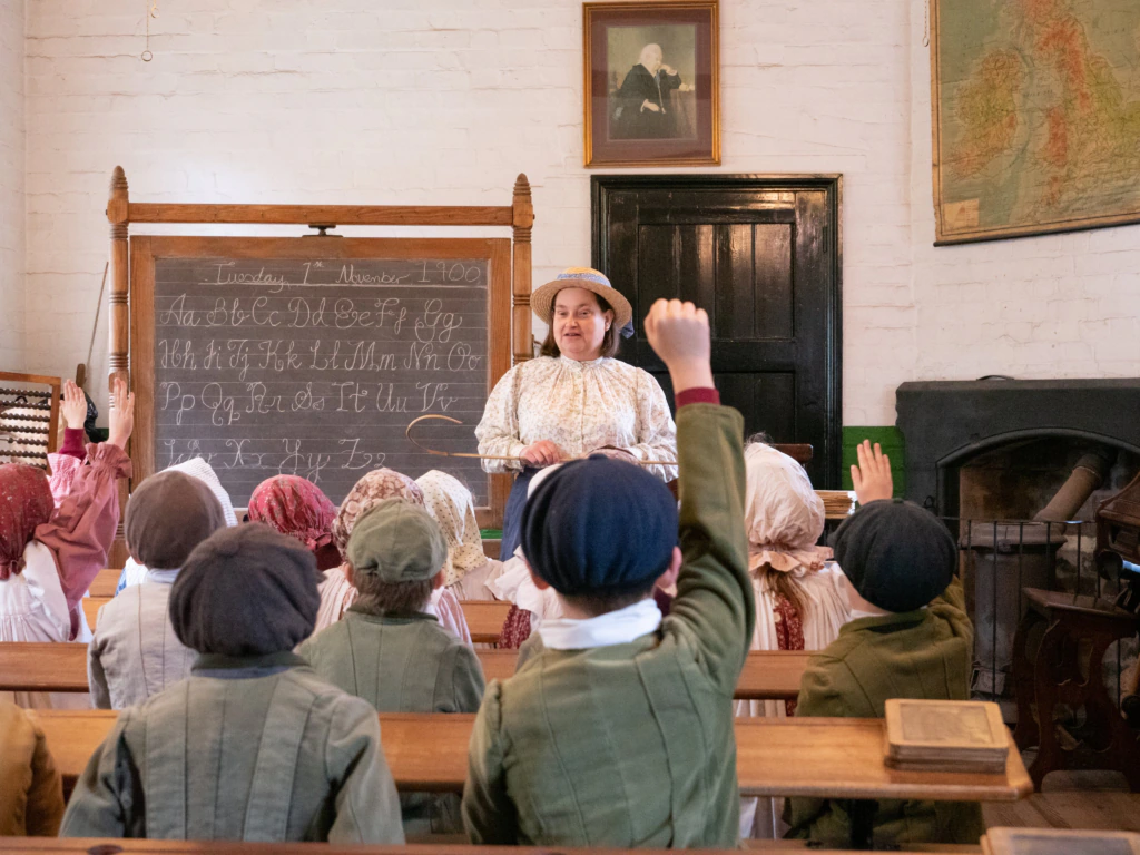 A woman in period clothing teaches young children, also in historical dress, in an old-fashioned classroom with a chalkboard, wooden desks, and hands raised to answer questions.