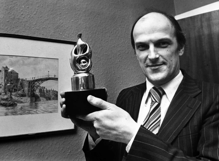 A man in a striped suit holds up a trophy with a stylized design, smiling at the camera. A framed picture of a bridge over water hangs on the wall behind him.