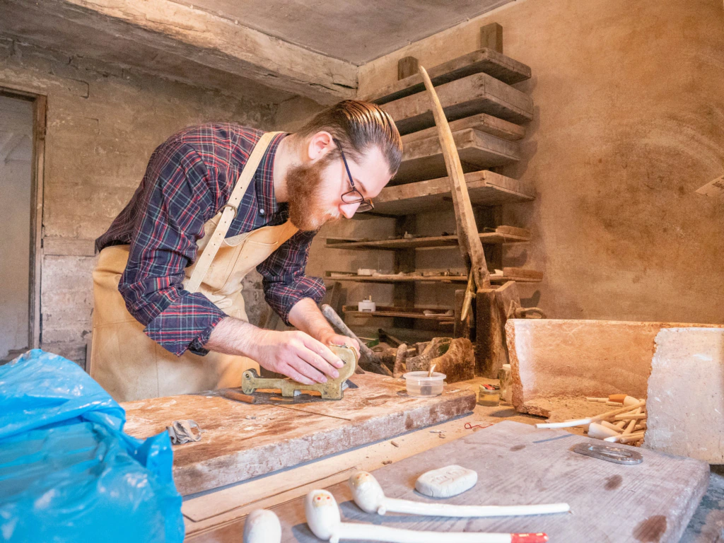 A man with a beard and glasses, wearing a plaid shirt and apron, works on a wooden project in a rustic workshop. Various tools and wooden pieces are scattered on the workbench. Shelves with materials are in the background.