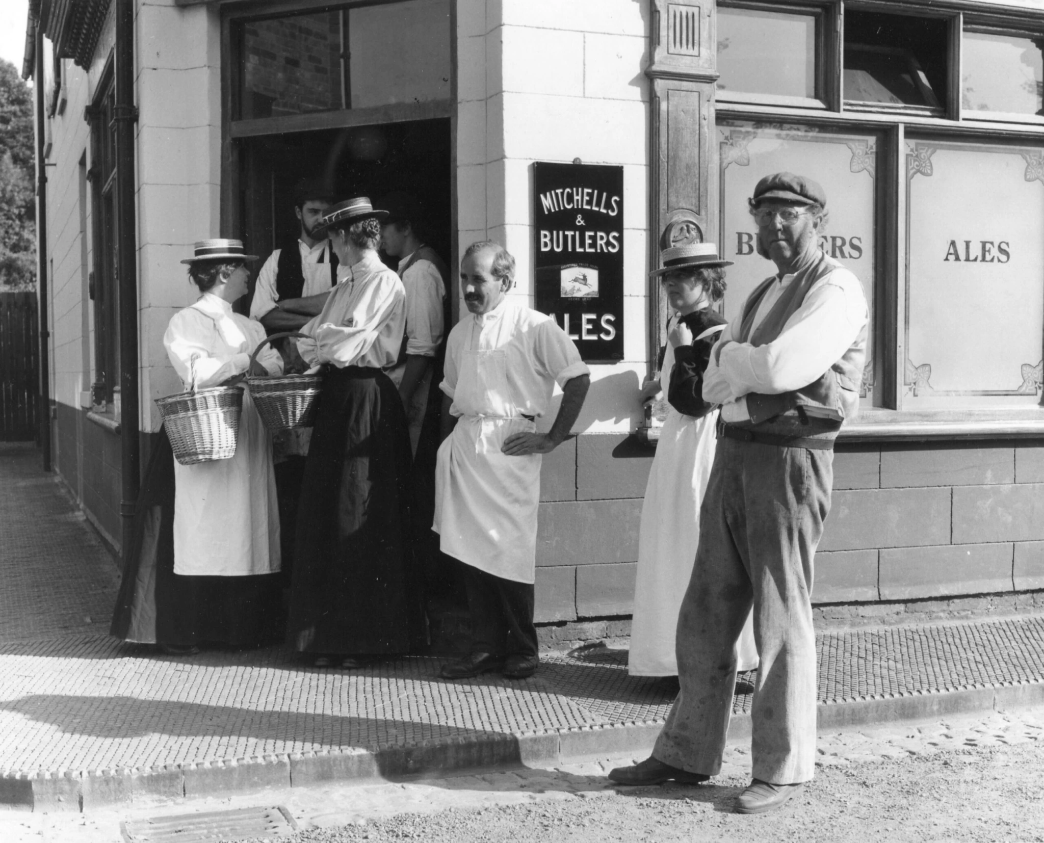 Black and white photo of people outside a pub, some in aprons and hats, with baskets; a sign reads “Mitchells & Butlers Ales.” Two men in caps stand in the foreground, while others gather by the entrance.