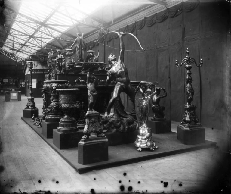 A black and white photo of an exhibition hall displaying ornate bronze sculptures, including a prominent statue of a nude archer drawing a bow, surrounded by candelabras and classical-style figures on pedestals.