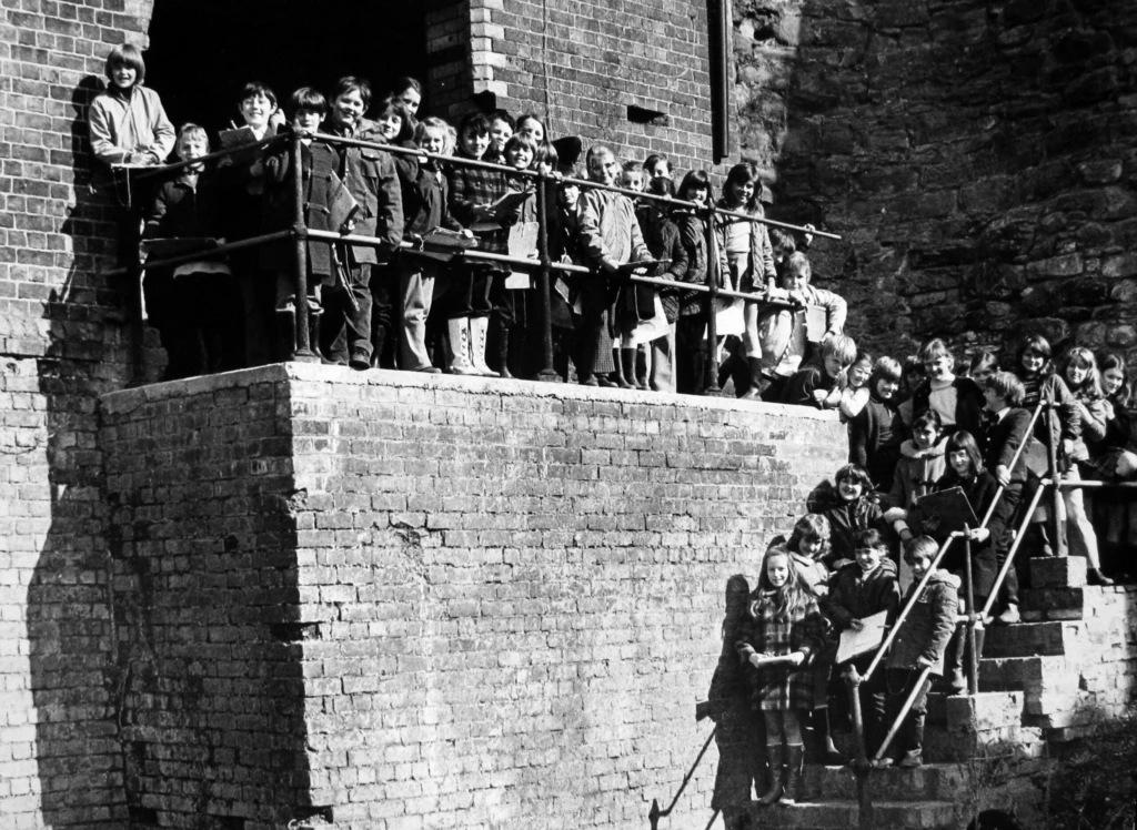 A large group of children stand on brick steps and a platform next to a stone wall, posing for a photo in an outdoor setting. The image is in black and white.