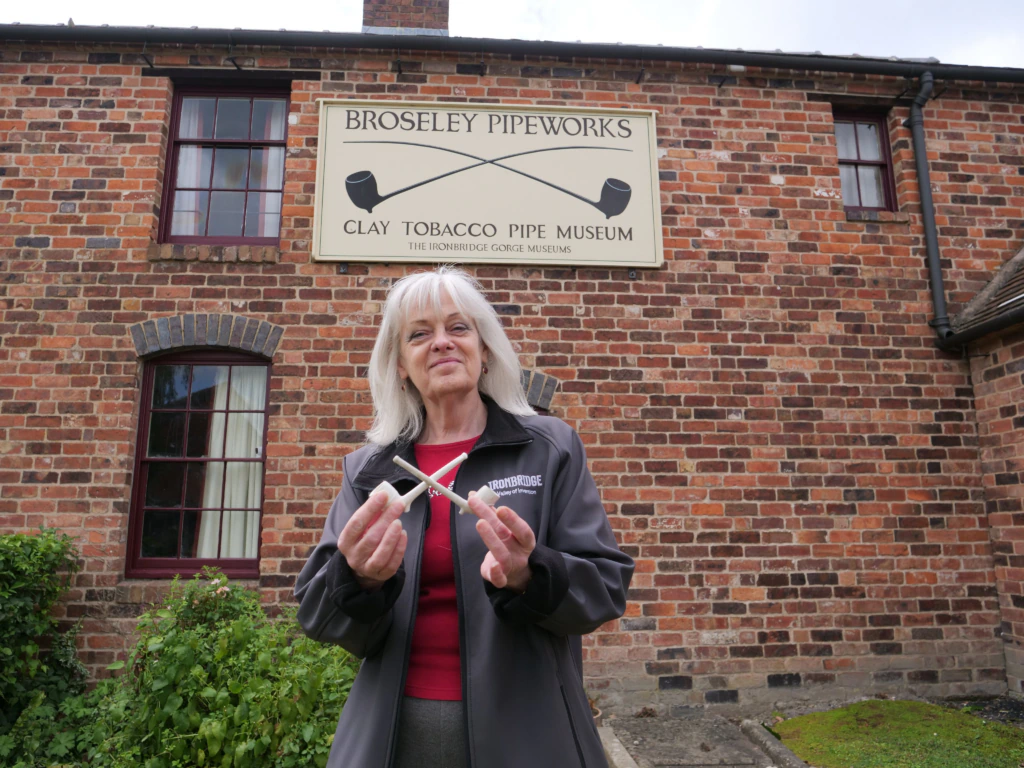 A woman stands in front of a brick building with a sign reading “Broseley Pipeworks Clay Tobacco Pipe Museum,” holding two clay pipes and smiling at the camera.