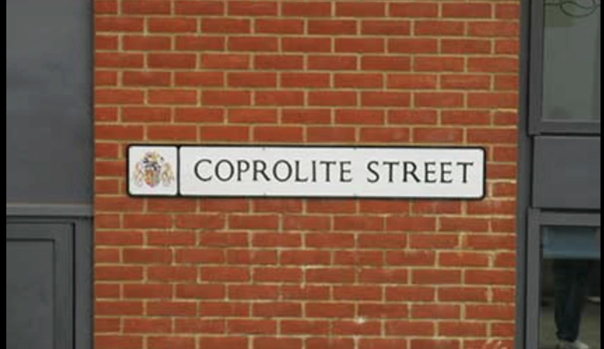 A street sign reading COPROLITE STREET mounted on a red brick wall, with parts of windows visible on either side.