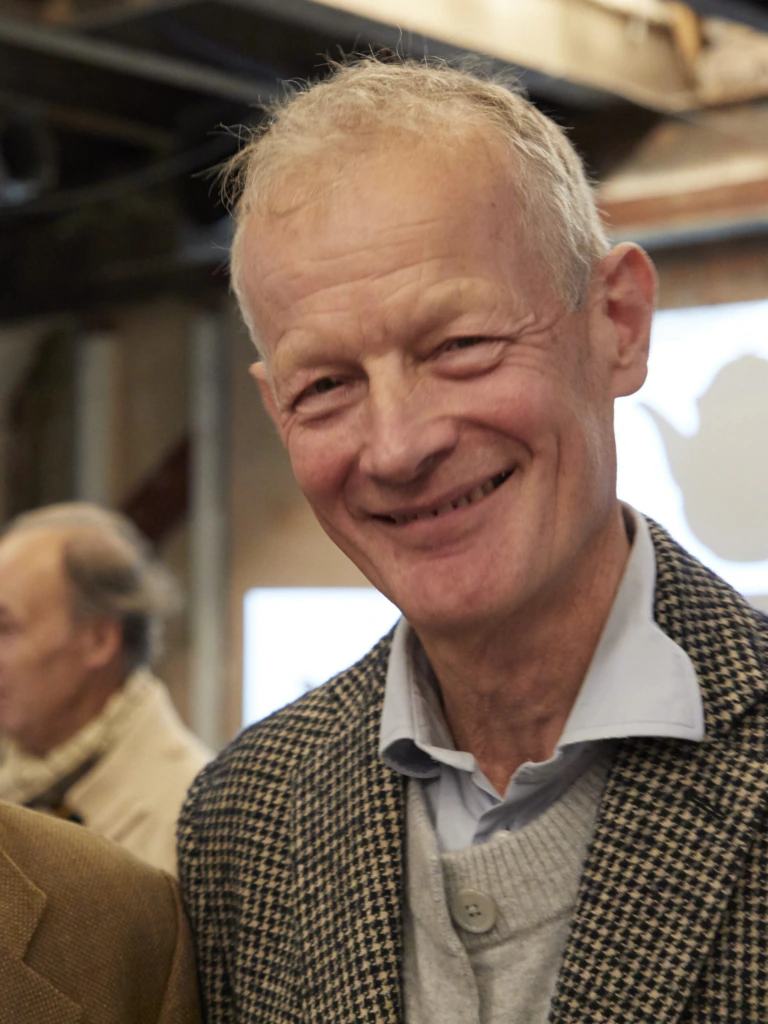 An older man with short gray hair is smiling, wearing a gray sweater, collared shirt, and checked blazer. The background is an indoor setting with another person and blurred details.
