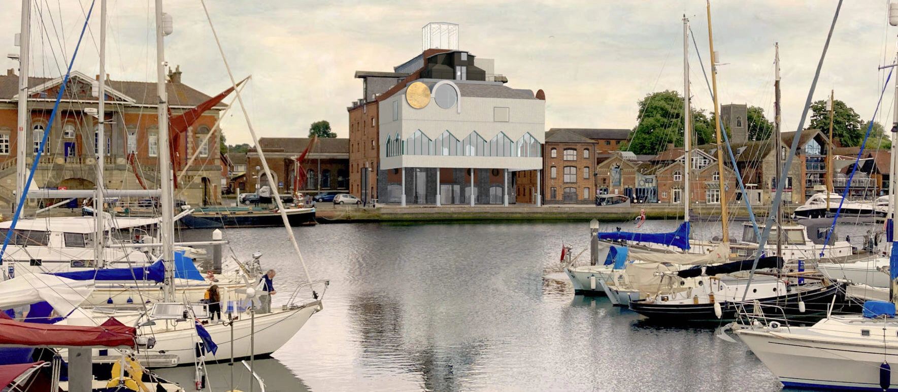 Sailboats are docked in a marina with calm water, surrounded by brick buildings. A large building with a clock and blue accents sits in the center, under a cloudy sky. People are visible on some of the boats and docks.