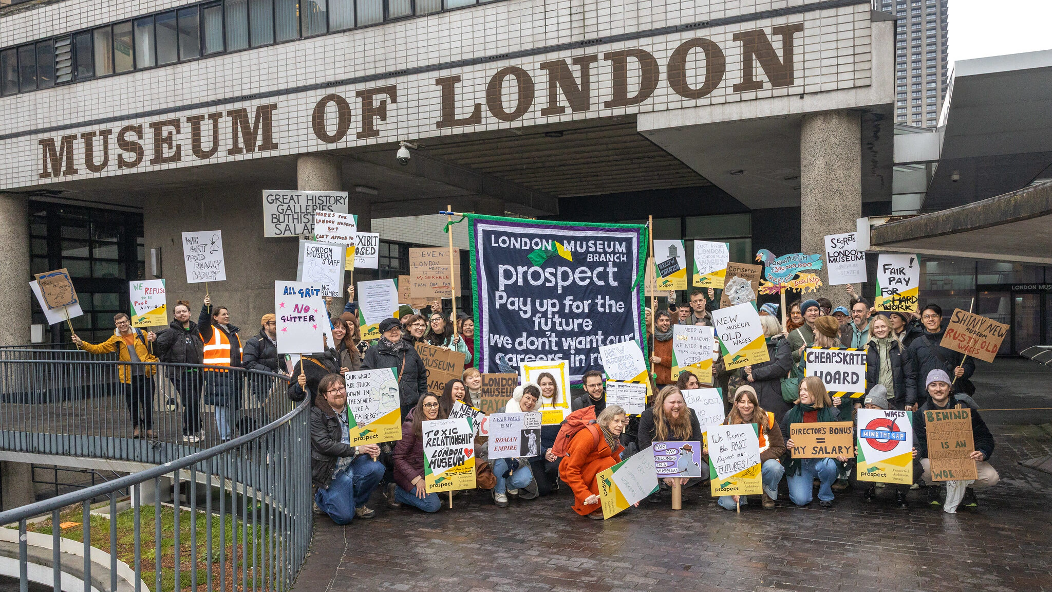 A large group of people protest outside the Museum of London, holding signs demanding fair pay and improved working conditions. Some signs read Pay up for the future, Support museum workers, and End gender pay gap.