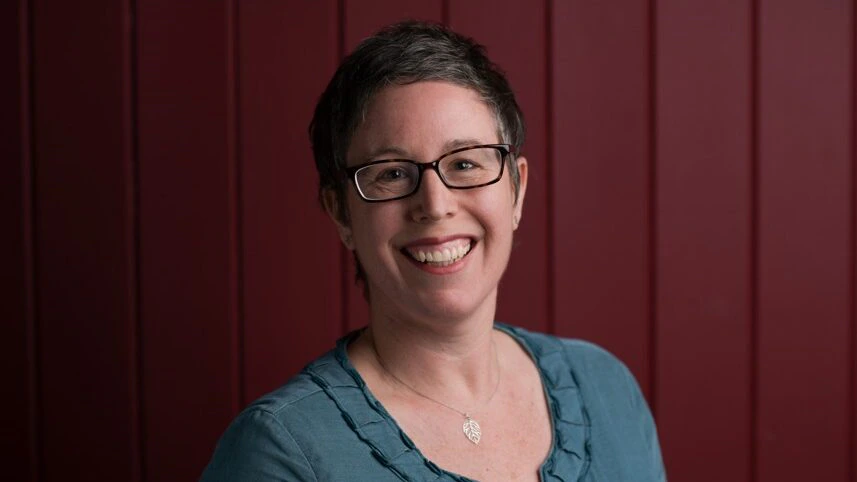 A person with short dark hair, glasses, and a blue ruffled shirt smiles at the camera. They wear a leaf-shaped pendant necklace and stand in front of a maroon, vertically paneled background.