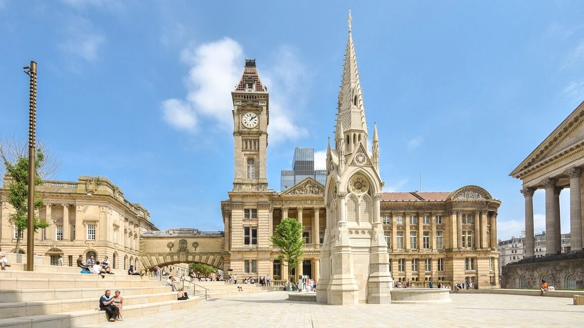 A sunny city square with a gothic-style clock tower, a spired monument in the foreground, and ornate historic buildings surrounding the open area where people are sitting on steps.