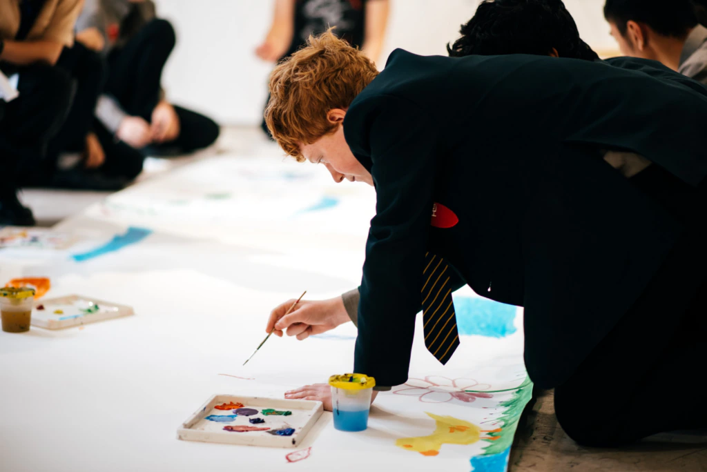 A student in a school uniform kneels on the floor, painting a colorful picture on a large sheet of paper with paint and brushes. Other students are in the background, also working on art projects.